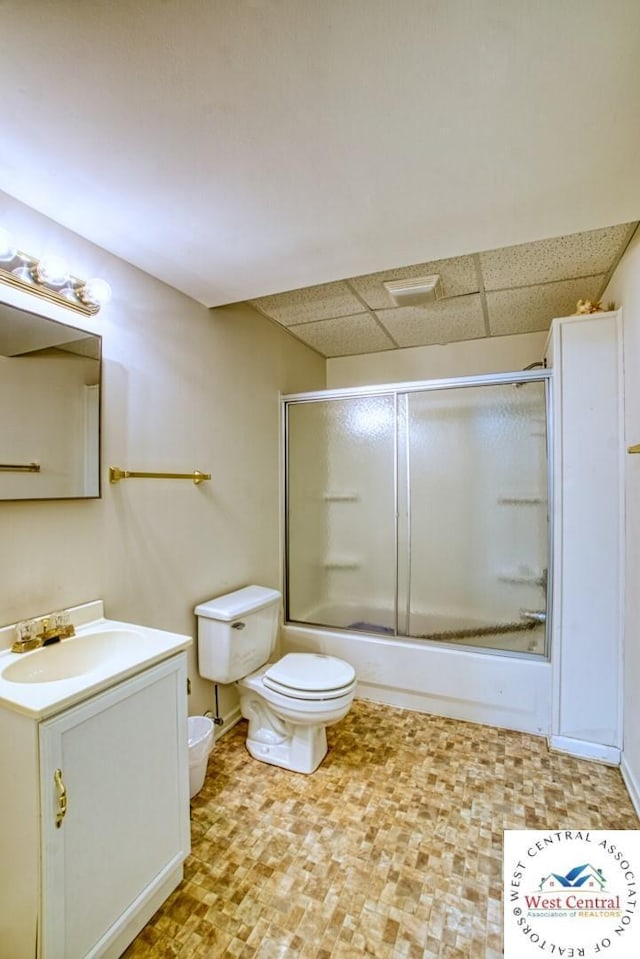 bathroom featuring a drop ceiling, combined bath / shower with glass door, toilet, vanity, and baseboards