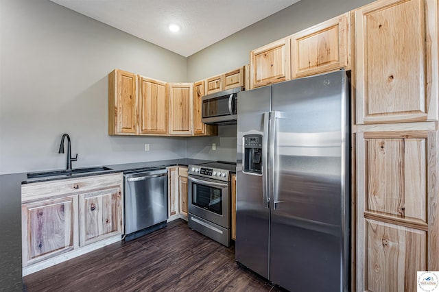 kitchen with light brown cabinetry, appliances with stainless steel finishes, dark countertops, a sink, and dark wood-type flooring