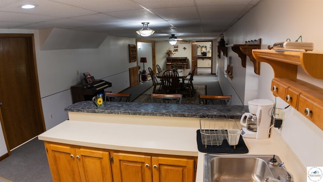 kitchen featuring brown cabinets, a sink, a ceiling fan, carpet, and a paneled ceiling