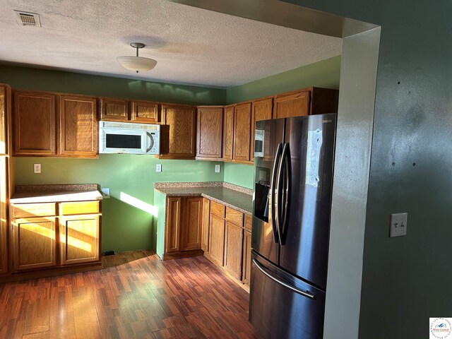 923 E Walnut St, Rich Hill, MO 64779 - kitchen featuring white microwave, brown cabinetry, dark wood-style flooring, stainless steel refrigerator with ice dispenser, and a textured ceiling