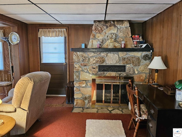 living area featuring a drop ceiling, a stone fireplace, wood walls, and carpet