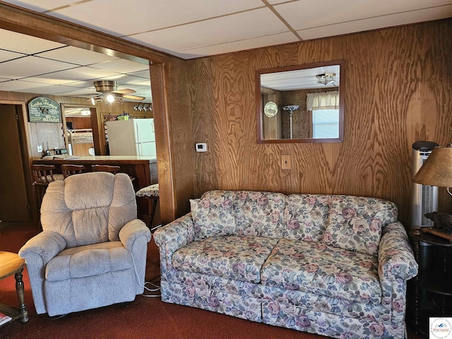 carpeted living area with ceiling fan, wooden walls, and a paneled ceiling