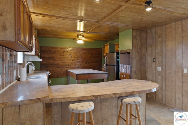 kitchen with wooden ceiling, freestanding refrigerator, wood walls, ceiling fan, and a sink