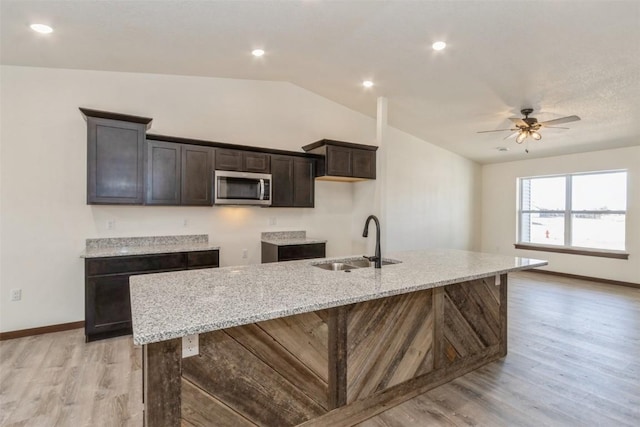 kitchen with a kitchen island with sink, a sink, stainless steel microwave, light wood-style floors, and lofted ceiling