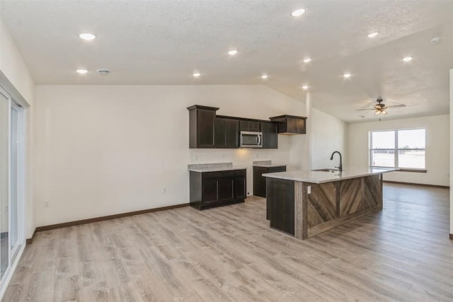 kitchen with lofted ceiling, an island with sink, a sink, stainless steel microwave, and light wood-type flooring