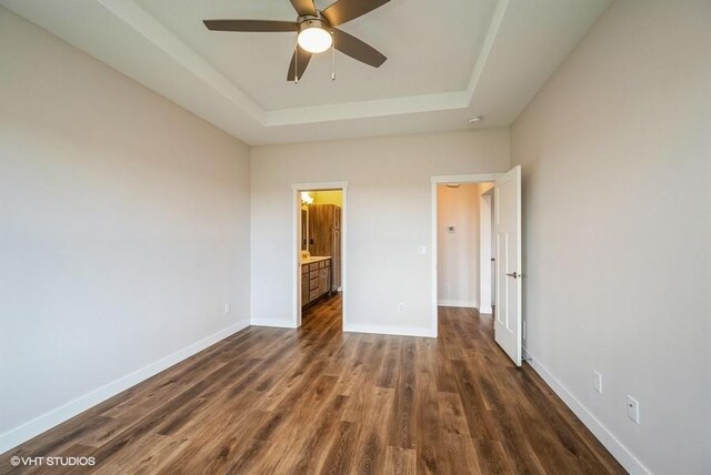 unfurnished bedroom with ceiling fan, a raised ceiling, connected bathroom, and dark wood-type flooring