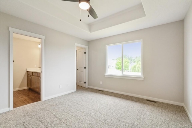 unfurnished bedroom featuring a raised ceiling, ensuite bath, ceiling fan, and carpet