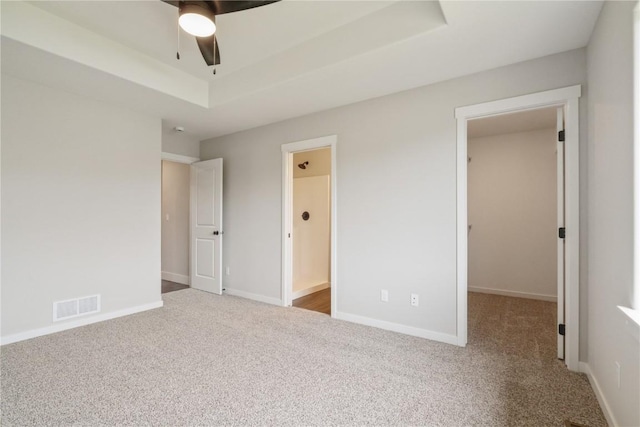 unfurnished bedroom featuring carpet flooring, a walk in closet, ensuite bath, a tray ceiling, and ceiling fan