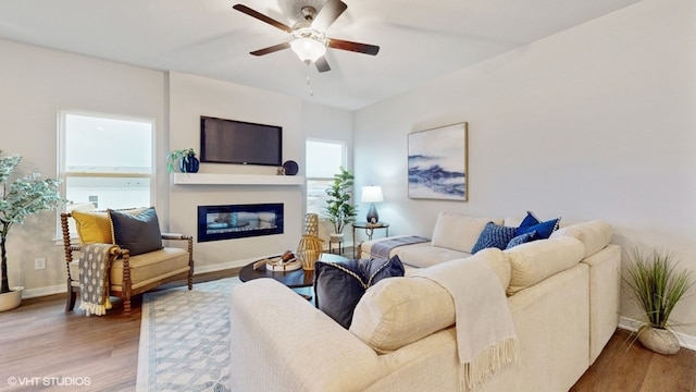 living area with a glass covered fireplace, baseboards, wood finished floors, and ceiling fan