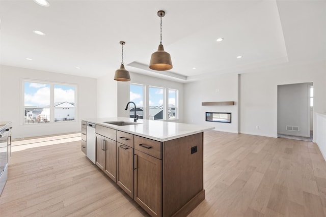 kitchen with an island with sink, sink, hanging light fixtures, a raised ceiling, and light wood-type flooring