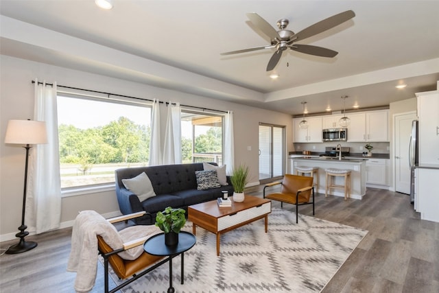 living room with a raised ceiling, ceiling fan, sink, and light wood-type flooring