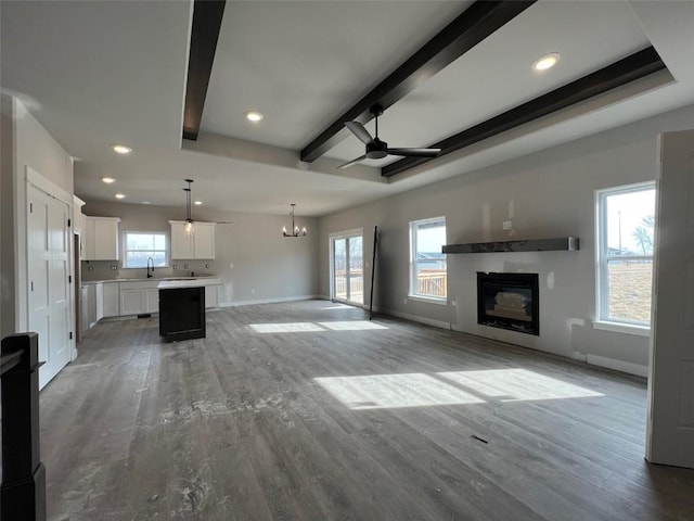 unfurnished living room with beam ceiling, ceiling fan with notable chandelier, a healthy amount of sunlight, and hardwood / wood-style flooring