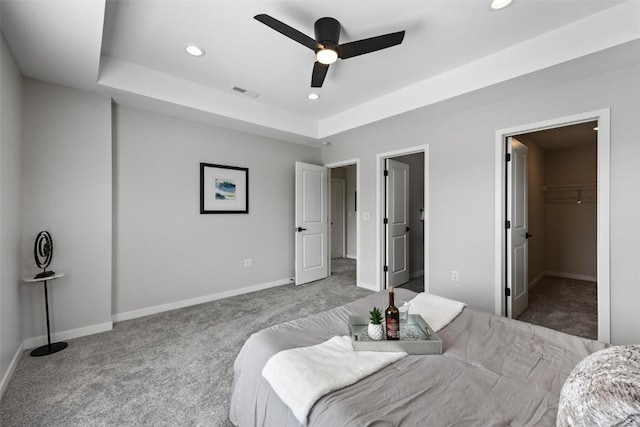 bedroom with ceiling fan, a walk in closet, a closet, a tray ceiling, and light carpet