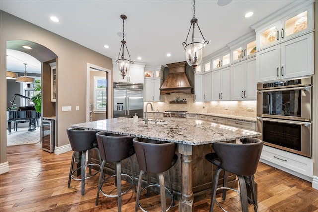 kitchen featuring hardwood / wood-style flooring, a kitchen island with sink, white cabinetry, custom exhaust hood, and stainless steel appliances