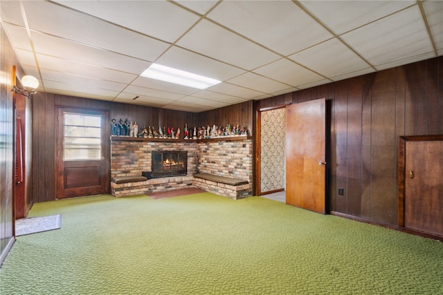 basement with a drop ceiling, a brick fireplace, wood walls, and carpet flooring