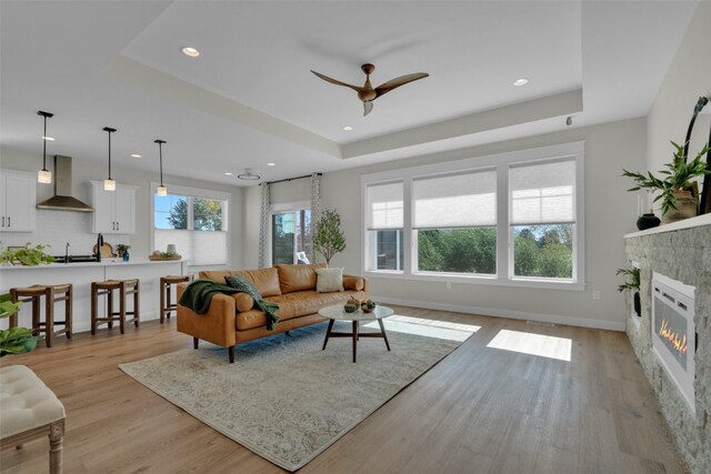 living room with ceiling fan, a raised ceiling, light hardwood / wood-style flooring, and a stone fireplace