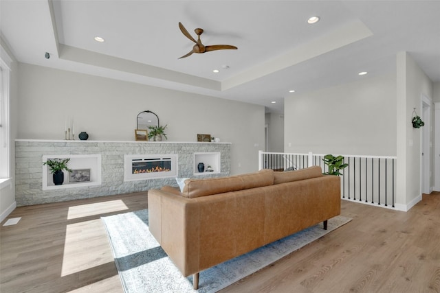 living room with light hardwood / wood-style flooring, a stone fireplace, a tray ceiling, and ceiling fan