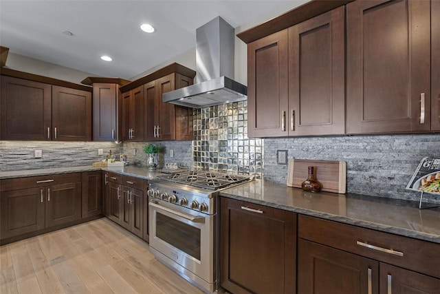 kitchen featuring wall chimney range hood, tasteful backsplash, high end stove, light hardwood / wood-style flooring, and dark stone countertops