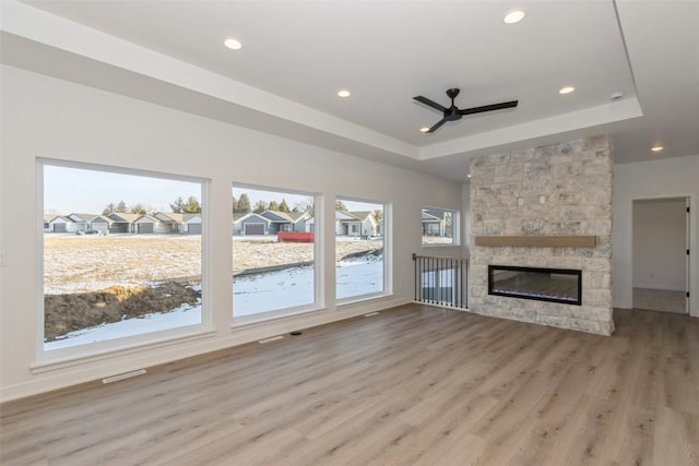 unfurnished living room featuring a raised ceiling, light hardwood / wood-style flooring, a stone fireplace, and ceiling fan