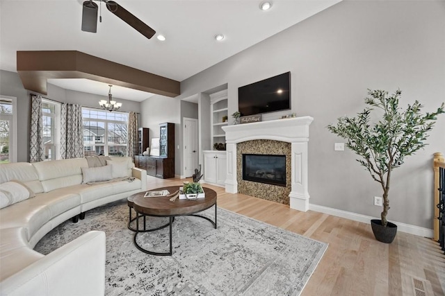 living room with built in shelves, ceiling fan with notable chandelier, a high end fireplace, and light hardwood / wood-style floors