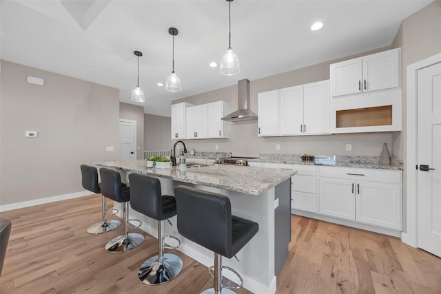 kitchen with white cabinets, a center island with sink, hanging light fixtures, light hardwood / wood-style flooring, and wall chimney exhaust hood