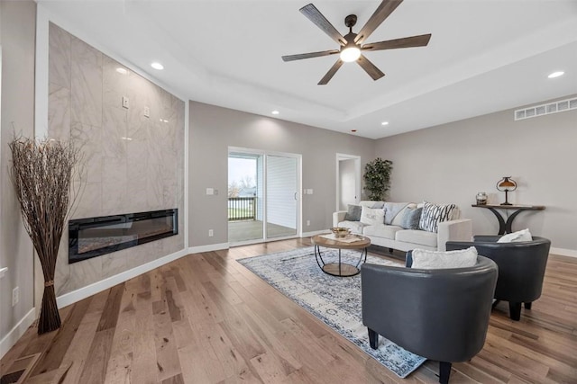 living room with a raised ceiling, a tile fireplace, ceiling fan, and light hardwood / wood-style flooring