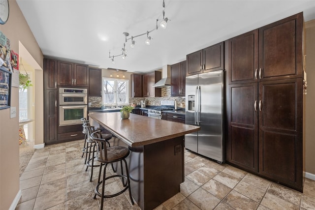 kitchen with a breakfast bar area, stainless steel appliances, wall chimney range hood, backsplash, and dark countertops