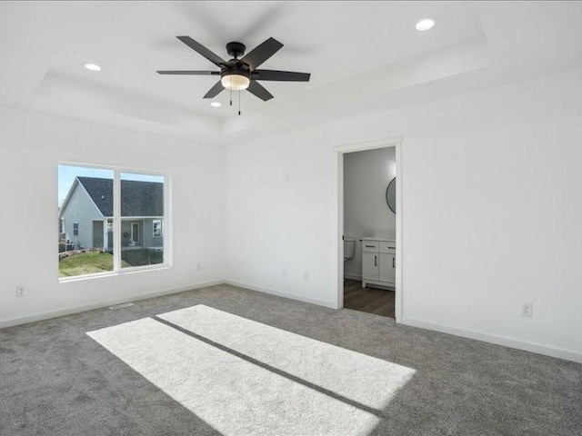 unfurnished bedroom featuring a raised ceiling, ceiling fan, ensuite bathroom, and dark carpet