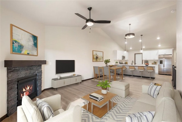 living room featuring a ceiling fan, a stone fireplace, light wood-type flooring, baseboards, and lofted ceiling