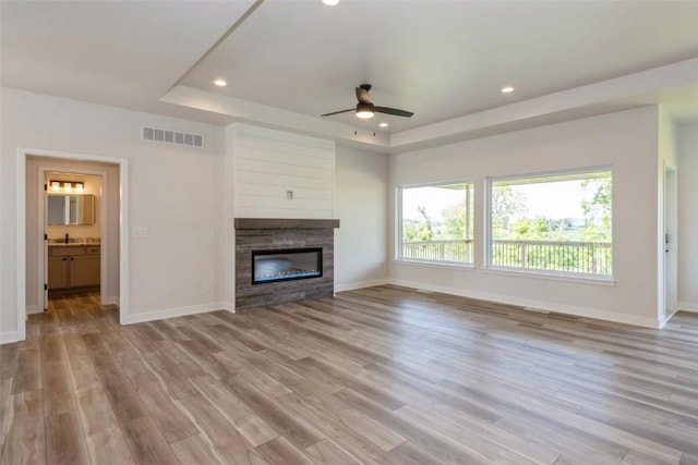 unfurnished living room featuring a tray ceiling, ceiling fan, a fireplace, and light wood-type flooring