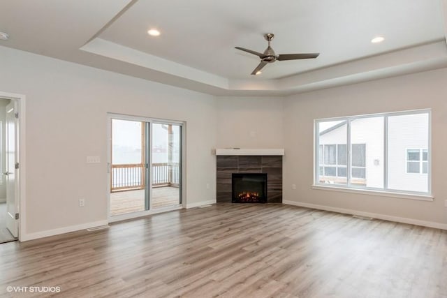 unfurnished living room featuring a tray ceiling, a tiled fireplace, ceiling fan, and light hardwood / wood-style floors