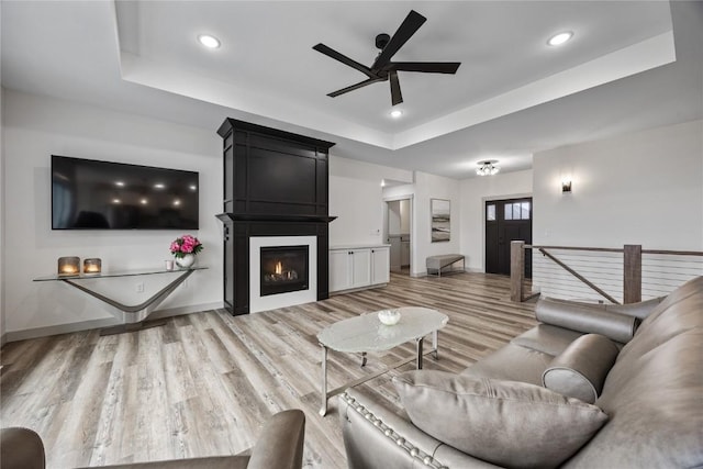 living room featuring a fireplace, recessed lighting, a tray ceiling, a ceiling fan, and light wood finished floors