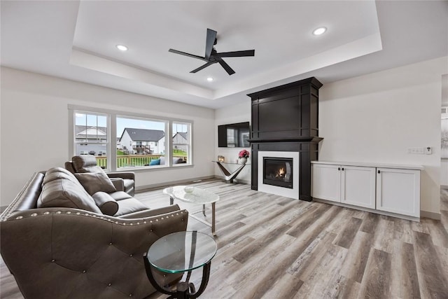living room featuring ceiling fan, a raised ceiling, a fireplace, recessed lighting, and light wood-type flooring
