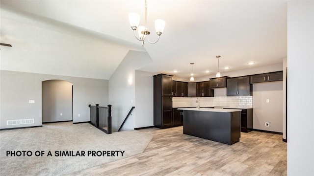 kitchen with dark brown cabinetry, vaulted ceiling, a kitchen island with sink, decorative light fixtures, and an inviting chandelier