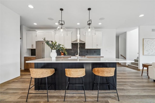 kitchen featuring light wood-type flooring, tasteful backsplash, wall chimney exhaust hood, white cabinets, and an island with sink