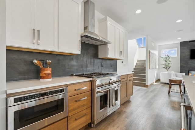 kitchen featuring backsplash, wall chimney range hood, hardwood / wood-style flooring, white cabinetry, and stainless steel appliances