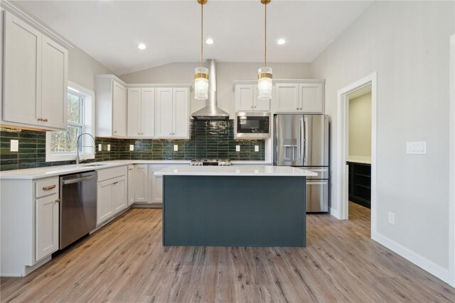 kitchen featuring wall chimney range hood, appliances with stainless steel finishes, decorative light fixtures, a kitchen island, and white cabinetry