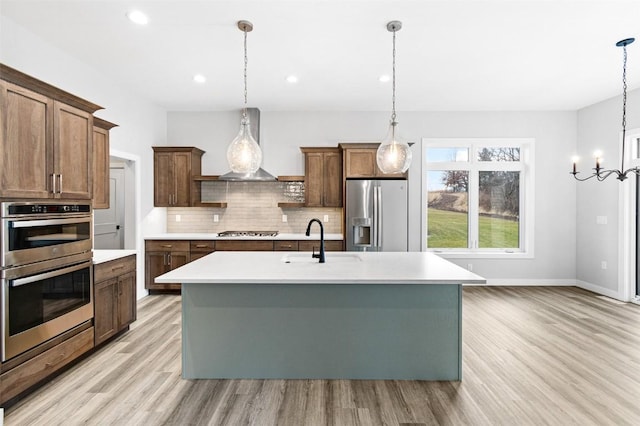 kitchen with stainless steel appliances, sink, wall chimney range hood, pendant lighting, and an island with sink