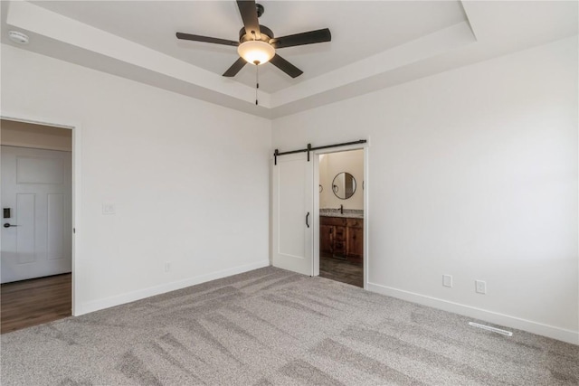 unfurnished bedroom featuring carpet, ensuite bath, ceiling fan, a barn door, and a tray ceiling