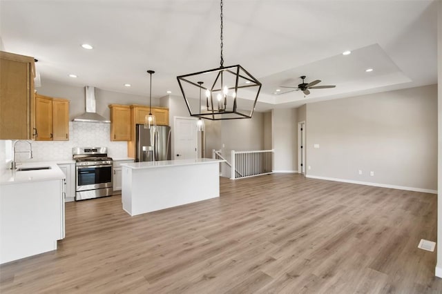 kitchen with wall chimney range hood, a tray ceiling, decorative light fixtures, a kitchen island, and stainless steel appliances