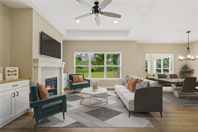 living room with ceiling fan with notable chandelier, light hardwood / wood-style floors, a raised ceiling, and a tiled fireplace