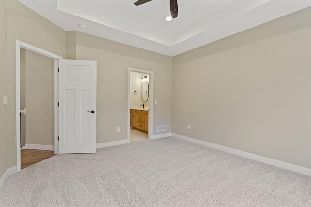 unfurnished bedroom with light carpet, a tray ceiling, ensuite bath, and ceiling fan