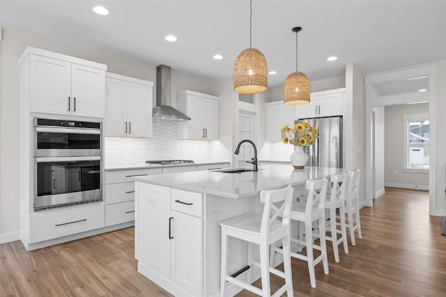 kitchen featuring a center island with sink, white cabinetry, wall chimney range hood, and appliances with stainless steel finishes