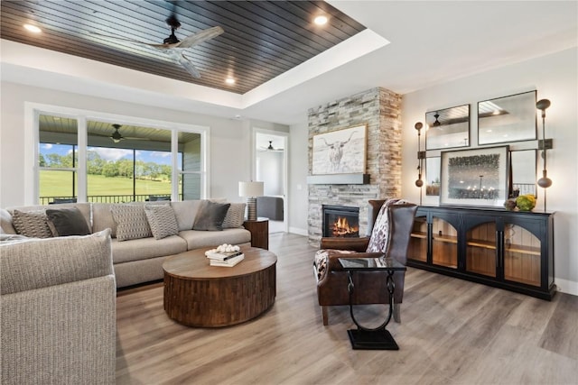 living room with a tray ceiling, ceiling fan, wood-type flooring, wooden ceiling, and a stone fireplace