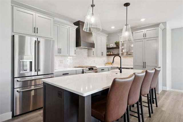 kitchen featuring an island with sink, stainless steel appliances, hanging light fixtures, and wall chimney range hood