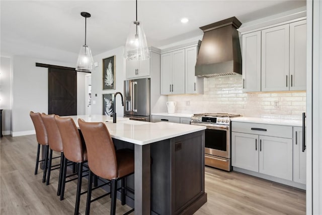 kitchen featuring premium range hood, stainless steel appliances, a kitchen island with sink, a barn door, and hanging light fixtures