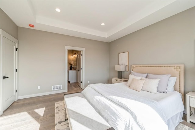 bedroom featuring light wood-type flooring, ensuite bathroom, and a raised ceiling