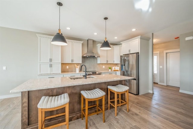 kitchen featuring wall chimney range hood, sink, stainless steel fridge with ice dispenser, a kitchen island with sink, and light stone counters