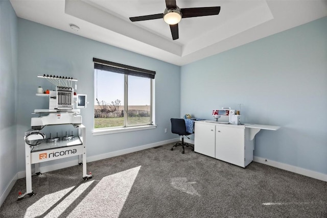 home office with ceiling fan, dark colored carpet, and a tray ceiling