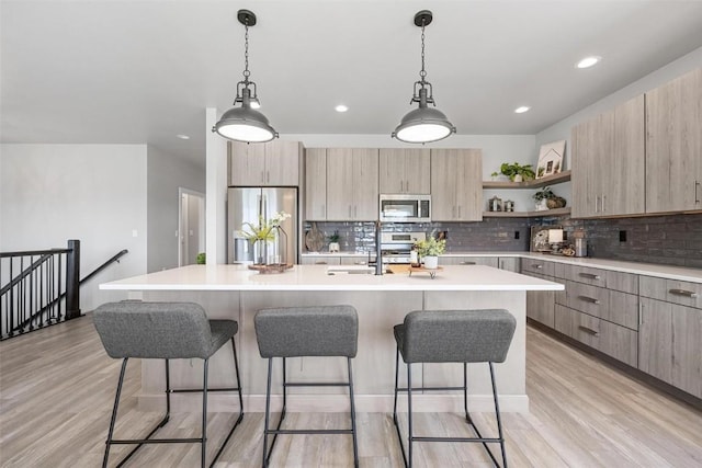 kitchen featuring appliances with stainless steel finishes, decorative light fixtures, an island with sink, backsplash, and light hardwood / wood-style floors